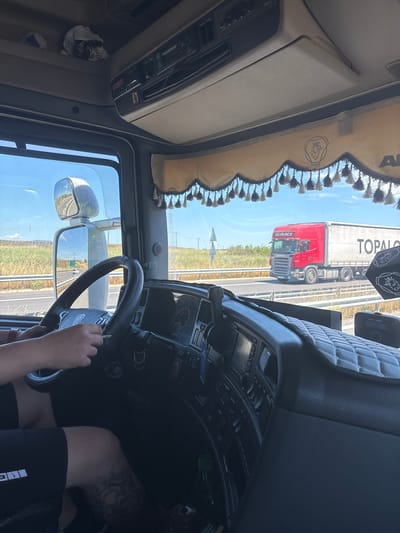 The image shows the interior of a truck cabin, looking out through the windshield. A person's hands are visible on the steering wheel, holding a cigarette. Outside the truck, a red semi-trailer truck is driving on a highway. The environment appears to be rural, with fields visible on either side of the road. The sky is blue and clear. The location is identified as Ardhani, Greece, implying the highway is likely part of the Greek road network near that area.