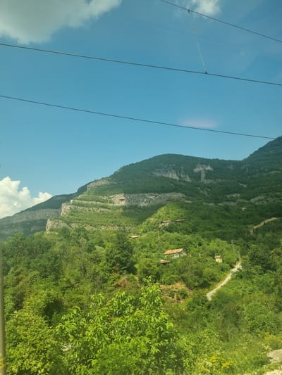 The image shows a verdant mountainous landscape under a clear blue sky with some scattered clouds. The terrain is characterized by steep, tree-covered slopes and rocky outcrops. Several houses with red-tiled roofs are visible nestled amongst the trees on the slopes, indicating a rural or semi-rural settlement. A winding road can be seen traversing the hillside, connecting the scattered dwellings. Power lines are strung across the sky in the upper portion of the image, and a pole is visible on the right side of the frame. The foliage is lush and green, suggesting a temperate climate during a warm season. The scene depicts a remote, picturesque setting, likely a village or hamlet situated within the natural beauty of the mountains. This scenery is typical of the region around Milanovo, Bulgaria, known for its mountainous terrain and scattered settlements.