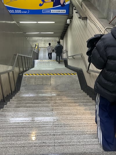 An overhead view captures a multi-tiered concrete and granite staircase descending into an underground passage in Yanqiao, China. In the foreground, the upper torso and legs of an individual wearing a black puffer jacket and navy blue sweatpants with white stripes on the side are partially visible on the right. Further down the central stairs, two individuals are seen from behind, descending away from the camera. The first wears a light-colored top and dark pants; the second, a darker top and dark pants.
The staircase features grey textured steps, with black and yellow striped hazard tape marking the edge of a landing midway down the visible descent. Tactile paving strips are present on the landings. Stainless steel handrails line both sides of the stairwells. The walls are a mix of metallic panels on the left and light-colored, possibly tiled or striped surfaces on the right. The flat ceiling is equipped with linear fluorescent light fixtures, illuminating the scene. A large blue and yellow banner with Chinese text and imagery hangs above the upper landing, advertising "APP用户" (APP user) and "找工作" (find a job). A white security camera is mounted high on the right wall. The environment appears to be an urban subway station or pedestrian underpass.