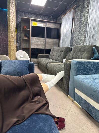 This image, captured in a furniture store in Barysaw, Belarus, shows a person's legs and feet resting on a blue sofa. The person is wearing white socks and dark red, pointed-toe shoes. In the background, there are other sofas, a large wardrobe with mirrored doors, and decorative walls with textured wallpaper. A fluffy white armchair is visible to the left of the wardrobe. The store appears well-lit with overhead fluorescent lighting.