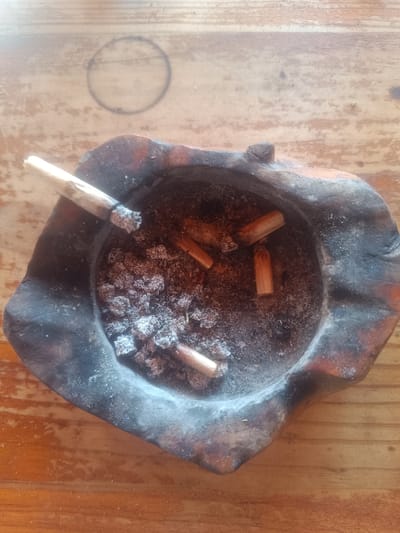 A dark, irregularly shaped wooden ashtray is centered on a light-colored wooden surface. The ashtray contains a mixture of grey ash, several small clumps of ash, and five light brown filter tips, consistent with used hand-rolled cigarettes. One partially consumed, hand-rolled cigarette, exhibiting an ash tip and a light brown filter, rests on the upper-left edge of the ashtray. The surrounding wooden surface displays a visible grain pattern and a faint, dark circular ring mark situated above the ashtray. No individuals or active interactions are depicted. The scene is illuminated by ambient light. This scene is located in Forest Hills, Spain.