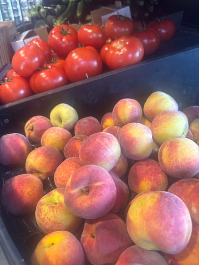 The image shows a close-up view of two black bins filled with produce. The bin on the left contains red tomatoes, while the bin on the right is overflowing with peaches. The location is a retail setting in an unknown location, but suggested to be in Marcq-en-Barœul, France. There are no people visible in the image. The primary objects are the tomatoes and peaches, arranged densely within their respective bins. The image lacks explicit actions or interactions. The environment appears to be a well-lit store, likely a supermarket or farmers market.