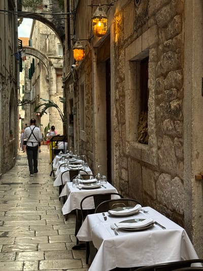 The image depicts an outdoor dining area with tables set for a meal. Two people are visible, one in the foreground wearing a white shirt and dark pants, facing away from the camera, and another person further back wearing a pink top and white pants. The scene appears to be a narrow street or alleyway, with stone architecture and arches suggesting a historic European city. The tables are covered with white tablecloths and set with silverware and folded napkins. Warm-toned lighting from industrial-style fixtures illuminates the area, hinting at either early morning or evening. The stone paving on the ground is worn and textured. The overall atmosphere suggests a charming, old-world setting for dining, possibly in Split, Croatia, given the architectural style. The weather appears to be fair.