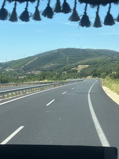 The image depicts a view from inside a vehicle looking out at a highway winding through a hilly, vegetated landscape. The road is asphalt with white lane markings. A metal guardrail runs along the left side of the road. In the background, a green, tree-covered hill rises, with power lines visible on its slopes. The sky is clear and blue. The foreground shows the interior of the vehicle, with decorative tassels hanging from the top edge of the windshield and a dark dashboard at the very bottom. The scene suggests a sunny day in a rural or semi-rural area. The location is specified as the city of Avra, Greece.