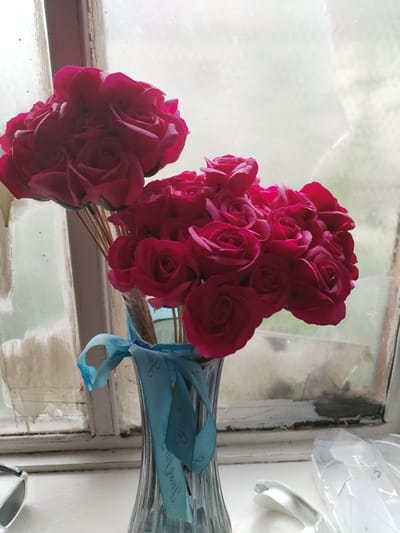 A bouquet of vibrant pink roses is displayed in a clear, fluted vase. The stems of the roses are tied together with a light blue ribbon that has text printed on it. The vase is placed on a white surface in front of a window with a distressed, aged frame. The window pane is covered in a hazy, translucent film, obscuring the view outside. To the left of the vase, a pair of sunglasses rests on the white surface. To the right, a white plastic case and utensil are visible. The scene is lit by soft, natural light filtering through the window. The location in the city of Muhametjan Tüymebaev, Kazakhstan, is an indoor setting, likely a windowsill.