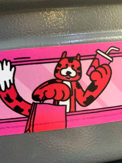 The image is a close-up of a promotional card featuring a cartoon tiger holding a shopping bag and a drink. The tiger is red with black spots and has a white chest and muzzle. The background is pink with diagonal stripes. Text in Ukrainian is visible at the bottom of the card. The card is placed on a textured grey surface. The location of Martynivka, Ukraine is not depicted in the image.