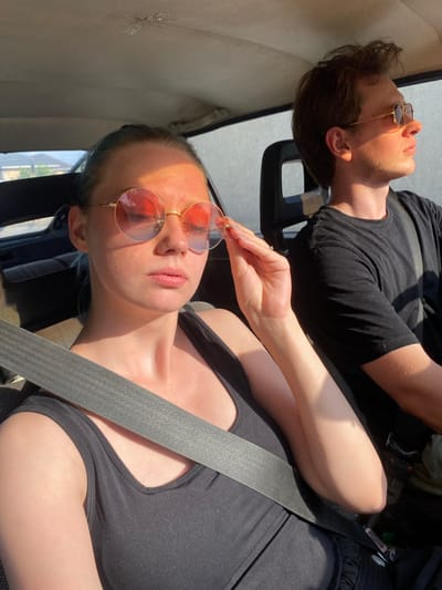 Two individuals, a woman and a man, are seated inside a vehicle. The woman, in the foreground, is wearing a black tank top and round, rose-tinted sunglasses. She is adjusting her sunglasses with her right hand, which is near her face. A gray seatbelt is visible across her chest. The man, in the background, is wearing a black t-shirt and sunglasses. He is looking out the window to his right. The interior of the car appears worn, with visible stitching and a slightly discolored headliner. Sunlight streams in, creating strong highlights and shadows on their faces and clothing. The background outside the car windows shows a residential area with houses and vegetation, consistent with the outskirts of a town or city. The context provided places this scene in the city of Lower Ala-Archa, Kyrgyzstan.