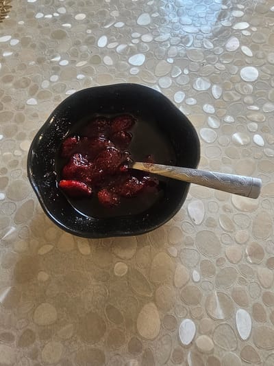 A black bowl filled with sliced strawberries in syrup sits on a patterned countertop. A silver spoon with an ornate handle rests inside the bowl, partially submerged in the syrup. The countertop has a pebble-like pattern with various shades of beige and white. The scene is lit from above, casting subtle shadows. The location in the city of Таёжное, Russia is not discernible from the image.