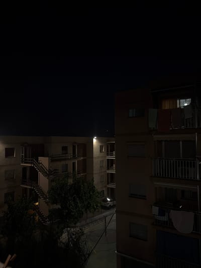 This is a nighttime photograph of an apartment building, with the image rotated 90 degrees counter-clockwise. The left side of the image is completely dark, suggesting the absence of light sources or features. The right side of the image shows the facade of a multi-story building.

On the upper floors, several balconies are visible. Some have clothes hanging out to dry, and one balcony has what appears to be a large white sheet or blanket draped over a railing. Metal security bars are present on some windows and balconies.

Below these upper floors, there are more windows and balconies. External staircases with metal railings are a prominent feature on the right side of the building, suggesting a fire escape or an alternative access route.

A large tree with dense foliage is positioned next to the building, its leaves illuminated by ambient light. Electrical wires are visible in the upper right portion of the frame.

The time of day is clearly night, indicated by the deep darkness and artificial lighting. No people are visible in the frame. The setting appears to be an urban residential area, likely in a warm climate given the clothing on the balconies. There is no visible text in the image. The overall mood is quiet and still, typical of a residential building at night.
