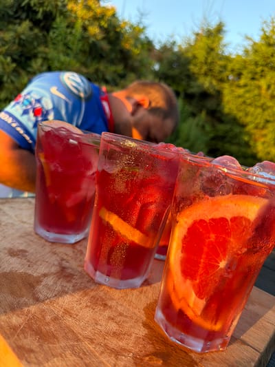 Three glasses of a red beverage, each containing ice and orange slices, are positioned on a wooden surface. A person, wearing a blue shirt with a white logo, is blurred in the background, lying down. The scene appears to be outdoors, with trees visible in the background and a clear sky above. The lighting suggests it is either sunrise or sunset. The specific location in Mordarka, Poland, cannot be determined from the image alone.
