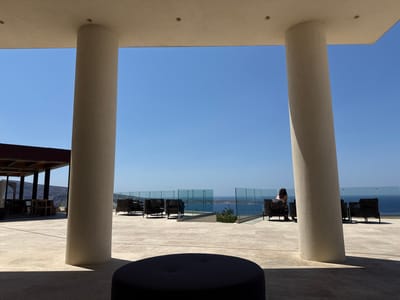 The image presents an elevated outdoor terrace overlooking the Aegean Sea, situated within the Ia Municipal Unit, Greece, under a clear blue sky. In the foreground, two large, light-colored cylindrical pillars, positioned symmetrically, frame the vista, casting shadows from overhead sunlight. A dark, circular, low-profile object, possibly an ottoman or table, is partially visible in shadow at the bottom center. The terrace floor is constructed from light-toned paving material.
Beyond the pillars, the sunlit terrace extends, furnished with multiple outdoor seating arrangements. To the left, a wooden pergola shades an area with several empty wooden tables and matching chairs. To the right, a collection of dark-colored, woven outdoor armchairs is visible. A transparent glass barrier with a dark top rail delineates the edge of the terrace from the expansive sea beyond. One individual, positioned on the right side of the terrace, is seated in a dark armchair, facing away from the viewer and towards the sea; the person has dark hair. The background consists of the calm, deep blue sea stretching to the horizon, where it meets a clear, light-blue sky. Distant landmasses are faintly visible on the left horizon. No discernible interactions or specific actions beyond being seated are depicted.