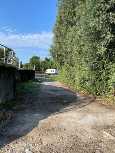 The image depicts an outdoor scene in Lokeren, Belgium, possibly near industrial or water management facilities. In the foreground, a dusty, uneven path made of dirt and gravel curves to the right. To the left, a large concrete structure with a metal railing is visible, suggesting it might be part of a reservoir, pumping station, or similar infrastructure. A shovel with a red handle rests against the concrete. Further back, a paved road leads towards a closed metal gate. Beyond the gate, a white van with the blue logo "GLS" is parked. The background is dominated by lush green trees and foliage. The sky is blue with scattered white clouds. The overall impression is one of a utilitarian outdoor area with elements of both industrial construction and natural vegetation.