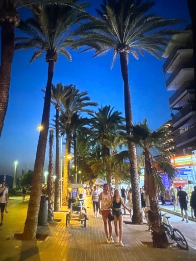 The image shows a street scene in Petarch, Bulgaria at night. Palm trees line the sidewalk, their trunks illuminated by streetlights. People are walking and cycling along the paved walkway. There are buildings with bright signage and lights in the background. Two young women are standing on the sidewalk, posing for a photo. A quadricycle is parked nearby.