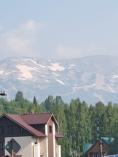 The image captures a panoramic view of a mountainous landscape with scattered patches of snow, likely in or near Sheregesh, Russia, a well-known ski resort. In the foreground, the roofs of several buildings, appearing to be houses or chalets, are visible. The architecture suggests a rustic style with dark tiled roofs and wooden elements. A dense forest of green trees separates the buildings from the imposing mountain range in the background. The sky is partly cloudy and a light blue. The overall scene conveys a sense of natural beauty and outdoor recreation, with hints of human settlement within a wilder environment. There are no people or animals visible in the image.