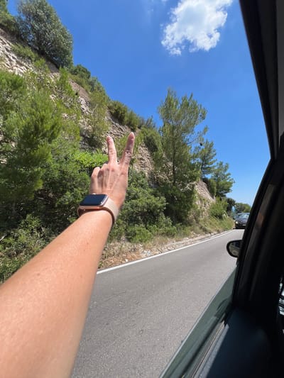 A first-person perspective shot shows a person's arm extended out of a car window, making a peace sign. The arm is tanned and has a light-colored smartwatch on the wrist. The car is driving on a paved road that curves to the right, with a steep, rocky hillside covered in green trees and bushes on the left. The sky is a clear, bright blue with a few white clouds. In the distance, another car is visible on the road. The location is the scenic coastal road near Mattinata, Italy, known for its dramatic cliffs and lush vegetation.