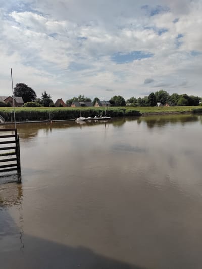 This is a photograph taken in Heiligenstedten, Germany, depicting a wide, muddy river in the foreground. The water's surface is calm and reflective, showing the sky and surrounding landscape. On the left side of the image, a dark wooden structure, possibly a pier or a dock, extends into the water. Several small white boats are moored along a distant embankment on the far side of the river. Behind the embankment, a row of houses with red-tiled roofs and green trees are visible under a cloudy sky. The overall impression is a serene, rural river scene.