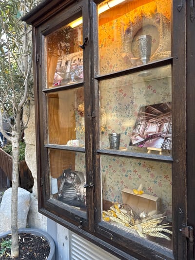 The image shows a close-up of a wooden display cabinet with glass doors, situated outdoors in Esslingen am Neckar, Germany. The cabinet is divided into four panes, with the left side showing a tree and some rocks outside, and the right side displaying various objects inside.

The top right pane features a wreath made of dried wheat and other natural materials, with a decorative metal cup placed in the center. The background of this pane is wallpaper with a floral pattern.

The bottom right pane contains a photograph of an indoor dining area with wooden tables and chairs, a low-ceilinged wooden roof, and a tiled floor. To the left of this photograph, on a wooden shelf, is another metal cup. The background is the same floral wallpaper. Further down, in the bottom right corner, is an open wooden box containing a bouquet of dried wheat and other decorative elements, with a small yellow butterfly figurine on top.

The left side of the cabinet displays items through the glass. The top left pane has a photograph of an archway and a staircase. The bottom left pane shows a vintage typewriter, and further down is another photograph of a staircase with archways.

The cabinet itself is made of dark wood and has metal hinges and latches. The lighting inside the cabinet appears warm, likely from artificial lights. The overall impression is of a quaint, decorative display, possibly of a shop or a historical establishment in Esslingen.