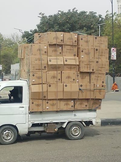 In Gyadi Gyadi Arewa, Nigeria, a white pickup truck is parked or moving on an asphalt road, heavily overloaded with numerous uniformly sized brown cardboard boxes. The boxes are stacked at least six layers high in the truck's bed, secured by multiple light-colored ropes or straps. Visible text on some boxes includes "Truck & Trak," "ELIMINATOR," "Doubt Busters?", and "ENCLOFMAC." The silhouette of a driver is visible within the vehicle's cab. To the right, on a concrete curb alongside the road, an individual dressed in an orange-brown full garment is seated on the ground, facing away from the truck, with a white bowl or object placed directly in front of them. The background features dense green foliage from trees, utility poles, and a pole displaying a red and white poster with a portrait. The sky is overcast.