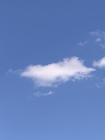 A single, elongated white cloud floats across a clear, bright blue sky. The cloud has a fluffy, somewhat wispy appearance, with subtle variations in its texture and shading. The sky is a uniform, vibrant blue, suggesting a daytime scene with good weather. The overall impression is one of calm and openness. There are no people, objects, or specific location cues visible in the image, beyond the cloud and sky. No text is present.