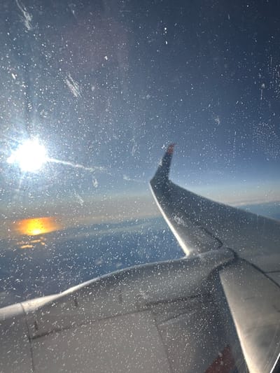 A distorted view from an airplane window reveals a wing extending across the right side of the frame. The wing's form is bathed in sunlight. Outside, the sky transitions from a deep blue at the top to a hazy horizon below. The sun shines intensely in the lower left corner. Numerous smudges and scratches cover the window, scattering the light and obscuring the view.