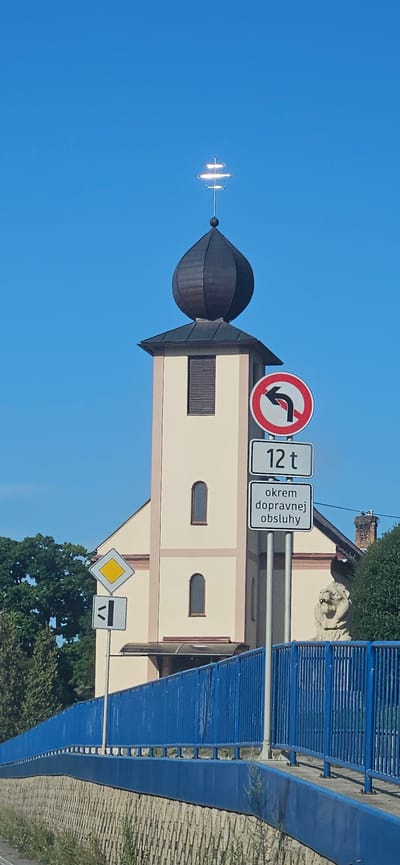 The image shows a church tower with a dark onion dome topped by a weather vane. In front of the tower, several traffic signs are visible: a red and white sign with a black arrow indicating no left turn, a rectangular sign displaying "12 t", and another rectangular sign with Slovak text "okrem dopravnej obsluhy". Below these, a yellow diamond-shaped sign with a black border is mounted, and beneath that is a white rectangular sign with black markings. A blue railing runs across the foreground, supported by a stone wall. A lion statue is partially visible to the right of the traffic signs. The scene is set against a clear blue sky with trees in the background and is identified as being in Giraltovce, Slovakia.