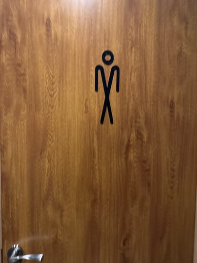 The image shows a wooden door with a black pictogram of a male figure, indicating a men's restroom. A silver-colored door handle is visible on the lower left side of the door. The door has a wood grain texture. The image was taken in Ruse, Bulgaria.