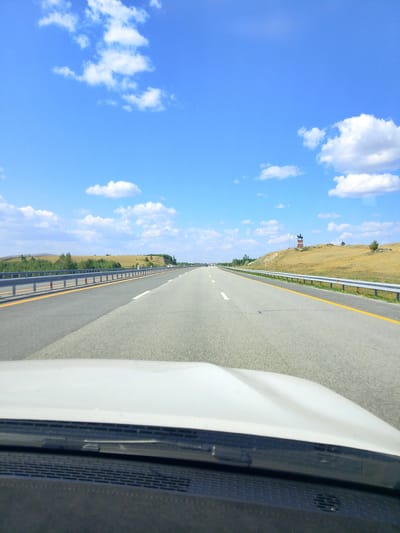 The image depicts a view from inside a vehicle driving on a highway. The highway stretches into the distance, with a bright blue sky and scattered white clouds overhead. On the right side of the road, a prominent monument stands on a hill. This monument appears to be a statue of a horse and rider on a pedestal, with a small structure resembling a watchtower or observation deck behind it. The surrounding landscape is characterized by rolling hills covered in dry, yellow grass, with scattered patches of green vegetation and trees. A guardrail runs along the side of the highway. The vehicle's dashboard and windshield are visible in the foreground.
