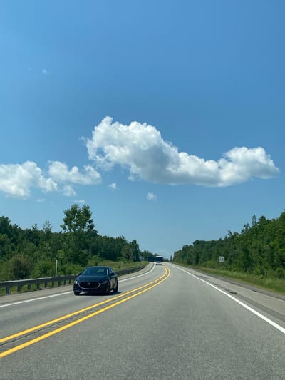 The image shows a paved highway in Pointe-au-Chêne, Canada, on a clear day with a bright blue sky and scattered white clouds. The road has two lanes in each direction, separated by double yellow lines. A black car is driving in the right lane, and a white bus is visible in the distance on the left lane. Lush green trees line both sides of the highway. A metal guardrail is present on the left side of the road, separating it from a grassy shoulder. The overall scene depicts a typical highway landscape during daytime travel.