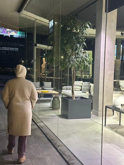 The image, rotated 90 degrees clockwise, presents an evening or nighttime view into a modern interior space, seen through a glass barrier. In the foreground, a person stands with their back to the viewer, observing the scene. They are dressed in a light beige, possibly quilted, long coat or jacket, a matching beanie, dark trousers, and light-colored slip-on shoes.

The interior, visible through the glass, features contemporary design elements. A large, leafy potted plant, resembling a eucalyptus tree, stands prominently on the left. To its right, furniture includes sofas with striped cushions and a dark, square cabinet or table. Further back, a lighter wooden table holds a yellow object and a green vase or jug. The architectural style incorporates grey, concrete-like pillars and extensive glass partitions, suggesting a commercial establishment like a café, lounge, or office lobby.

Reflections in the glass barrier on the lower left reveal text from a screen or sign. Clearly visible is "DR. ANGELOS KARATZIAS" followed by "ARISTIDIS THEOCLITOU & DEMETRIOS YIOGALLIS." Below this, the phrase "SECOND CUP" is present. There are also elements that appear to be from a sports broadcast, including the word "SPORTLIGHT" and some numerical scores like "23." Another small digital display behind the plant shows the number "88." The general atmosphere is calm, lit by artificial indoor lighting.
