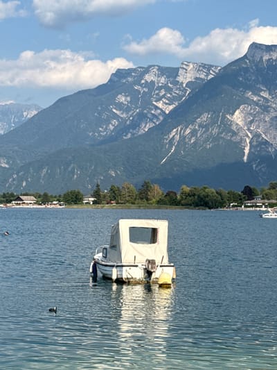 A motorized boat with a beige canopy is anchored in the blue water of a lake, with its reflection visible below. In the foreground, a black and white duck swims in the water. The middle ground shows a shoreline with buildings, trees, and a few people visible on the beach. In the background, a range of tall, rugged mountains with green slopes and rocky peaks dominate the scene under a bright blue sky with scattered white clouds. This scene is likely in Calceranica al Lago, Italy, given the presence of a lake and mountains typical of the region.