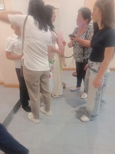 In Nor Hachin, Armenia, a group of individuals are gathered indoors, possibly at an event or gathering. The lighting is ambient and appears to be artificial. A young boy in a white collared shirt stands to the left, partially obscured by a woman wearing a white t-shirt and patterned pants. Beside her, another woman in a light-colored dress is visible, holding what appears to be a bouquet of flowers. Further to the right, an older woman in a patterned blouse is looking at a mobile phone. Next to her, a young woman in a dark t-shirt and light-washed baggy jeans is also present. The floor is tiled, and a rope barrier is visible in the background, suggesting a controlled or designated area. The overall atmosphere is informal and appears to be a social occasion.