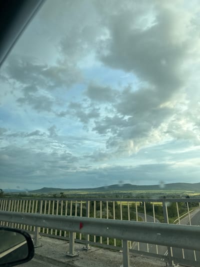 The image shows a view from a vehicle, likely a car, driving across a bridge in Kipra, Bulgaria. The sky is overcast with heavy clouds. To the right, a metal safety railing lines the bridge. Beyond the railing, a rural landscape unfolds with fields and forested hills. The road below is visible through the railing. The angle of the image suggests the photograph was taken from the passenger seat. A side mirror is partially visible in the lower right corner of the frame.