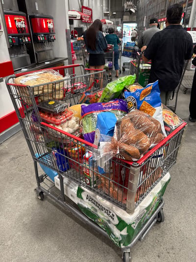 In the City of Yonkers, United States of America, a grocery shopping cart is filled with various items. The cart is positioned on a concrete floor, with a Coca-Cola soda fountain visible in the background. A woman in a denim jacket and black skirt is standing to the left of the cart, facing away from the camera. The cart contains a bag of bread, a container of avocados, a container of cherry tomatoes, a bag of pretzel crisps, a bag of pretzels, a package of hot dogs, a package of lettuce, and a package of toilet paper. Other shoppers and shopping carts are present in the background.