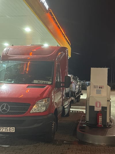 The image, taken at night in what appears to be a gas station or commercial area in Arad, Romania, shows a red Mercedes-Benz Sprinter-type van parked next to a fuel pump. The van is the primary subject, with its front end, including the grille with the Mercedes-Benz logo, headlights, and part of the bumper, prominently visible. The license plate, "7715 EO," is visible on the front bumper. On the windshield or dashboard, a sticker or sign reads "Krasi" with "BG" below it, indicating Bulgarian origin. Reflected on the van's window, the word "Volvo" can be seen, likely from an external source.
To the right of the van, a fuel pump station is visible. It is a large, white and black unit with a red fire extinguisher labeled "EXTINTOR" attached to its side. Several safety symbols (e.g., prohibition circles) are present on the pump's surface. In the background, illuminated by artificial lights, a building with a sloped, yellow-striped roof and a white wall is present. Part of a lit sign, reading "partener," is visible on the building's facade. The ground is paved with interlocking bricks, appearing somewhat wet, reflecting the artificial lights. The sky above is dark and clear. The overall scene suggests the van is either refueling or parked at a service station.