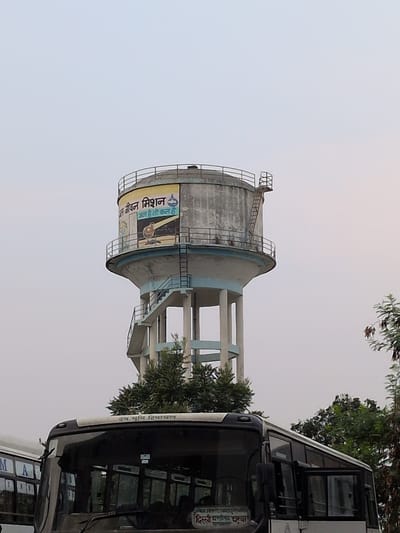 This is an outdoor, slightly tilted, vertical medium shot of a large, cylindrical water tower in Una, India. The sky is overcast and light, suggesting it might be dawn, dusk, or a cloudy day. The water tower is constructed from concrete and has a light blue and white paint scheme. It features a metal cage-like structure on top and a ladder leading up its side. A large, yellow banner is affixed to the tower's cylindrical body.

To the right of the water tower, a white bus is partially visible. The bus appears to be in motion or parked. The image captures a glimpse of the bus's front windshield, side mirrors, and part of its body, which is white with some dark trim.

The banner on the water tower contains text. The visible text on the banner is in Hindi script and reads: "स्वच्छ भारत जीवन मिशन" (Swachh Bharat Jeevan Mission) and below that "जल है तो जीवन है" (Jal hai toh jeevan hai), which translates to "If there is water, there is life." This indicates a public health or sanitation campaign related to water.

The text on the bus is also in Hindi and reads "देव भूमि हिमाचल" (Dev Bhoomi Himachal), meaning "Land of Gods Himachal." This likely refers to Himachal Pradesh, a state in India, suggesting the bus is operating in or from that region.

The overall scene is one of urban infrastructure combined with a public awareness campaign, set against a backdrop of a cloudy sky and greenery from trees. The orientation of the image suggests it may have been taken while moving or from an unusual vantage point.