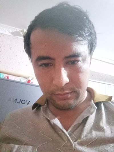 A medium close-up captures a man with dark hair receding slightly at the temples. He has dark eyes, a prominent nose, and a slight stubble. He is wearing a light-colored shirt with a subtle pattern and a contrasting, darker-colored collar. The background is softly lit, revealing a glimpse of a patterned wall and a piece of furniture with the word "VOLVO" mirrored on its surface. The lighting seems to be coming from above.
