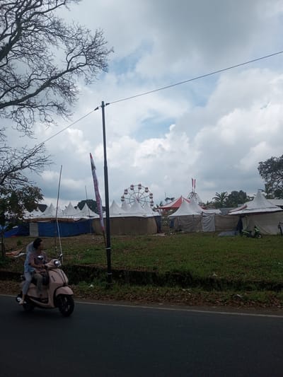 The image shows an outdoor event taking place in the city of Ban Phouviengnoy, Laos, marked by numerous white tents and a Ferris wheel in the background. In the foreground, a person is riding a pink scooter on an asphalt road, with another person as a passenger. A utility pole stands along the roadside. The sky is cloudy, and bare tree branches are visible in the upper left corner of the frame. The area between the road and the tents is a grassy field.