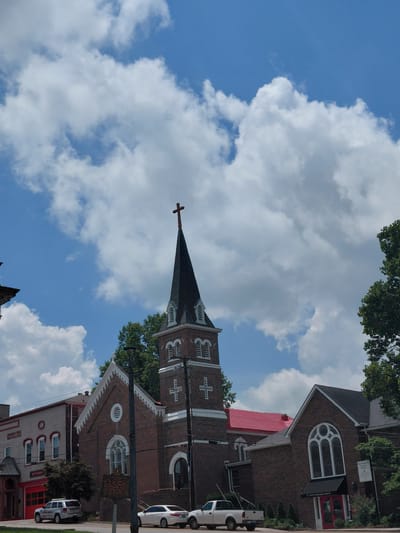 In Paris, Kentucky, USA, a brick church with a dark steeple topped by a cross stands against a bright blue sky filled with white, fluffy clouds. The church has arched windows and white trim. To the left of the church, a red-doored building with multiple windows is visible. Several cars are parked along the street in front of the buildings, including a white SUV with "FIRE DEPT" written on it. Trees are present on the right side of the image and behind the church. A historical marker is placed on a pole in front of the church.