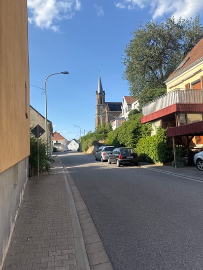 The image shows a street scene in Hüffler, Germany. On the left, a tan-colored building with a textured facade is visible. Adjacent to it is a paved sidewalk made of interlocking bricks. A metal street lamp with a modern fixture is positioned above the sidewalk. Further down the street, several parked cars are visible, including a black hatchback with a license plate reading "KUS 1 CS 75". On the right side of the street, a hill is covered with lush green vegetation. Several houses are nestled on this hillside, featuring red-tiled roofs and balconies. A prominent church spire, characteristic of German architecture, rises above the trees, indicating a church is nearby. The sky is a clear blue with scattered white clouds. The overall impression is a quiet residential street in a European town.