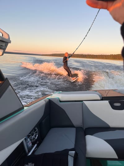A person in a black wetsuit is wakeboarding on a calm body of water, illuminated by the warm glow of sunset. The person is holding onto a tow rope connected to an unseen boat. The wakeboarder is positioned on the right side of the frame, creating a spray of water. In the foreground, the interior of a boat is visible, with upholstered seats in shades of gray, black, and white. A bottle of water and a speaker are also present in the boat. The background features a treeline along the horizon, with a gradient sky transitioning from pale blue to orange. The scene appears to be taking place on Lake Gromovskoye in Gromovo, Russia.