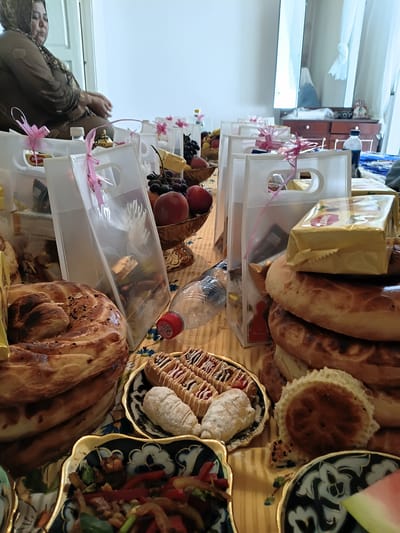 The image depicts a close-up, eye-level view of a table laden with food and gift bags, likely set up for a gathering or celebration. In the background, a woman wearing a headscarf and a traditional patterned dress is partially visible, seated and looking down. The table is covered with a yellow and white striped cloth.
Dominating the foreground are various baked goods, including large, round, braided breads with sesame seeds and smaller, flatter bread pieces. A decorative plate holds several pastries, some topped with icing and jam, and others appear to be crescent-shaped and dusted with powdered sugar. Another ornate plate in the lower left corner contains a colorful dish of stir-fried vegetables with meat. To the right, a slice of watermelon is visible on a dark patterned plate.
Scattered throughout the scene are several clear plastic gift bags, many tied with pink ribbon. These bags contain various items, including what appear to be chocolates, candies, and possibly other small gifts. A plastic water bottle with a red cap is positioned in the center of the frame. In the middle ground, a bowl filled with grapes and peaches is also visible. The overall impression is one of abundance and preparation for an event. The image appears to have been taken indoors, with natural light filtering in from a window or doorway on the left, illuminating the scene. The presence of a dresser with decorative items and curtains in the background suggests an indoor domestic setting, possibly in the city of Osh, Kyrgyzstan, given the cultural context hinted at by the woman's attire and the food items.