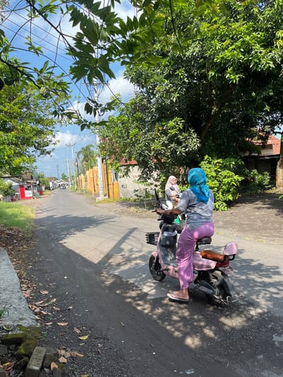 In Jati Alun Alun, East Java, a woman wearing a blue headscarf and grey shirt is riding a pink electric scooter down a paved road. She is wearing purple pants and white sandals. Another woman wearing a light-colored headscarf and grey shirt is a passenger on the scooter. The scooter has a basket on the front and a luggage rack on the back. The road is lined with trees and houses. The sky is blue with some clouds. The scene appears to be a residential street on a sunny day.