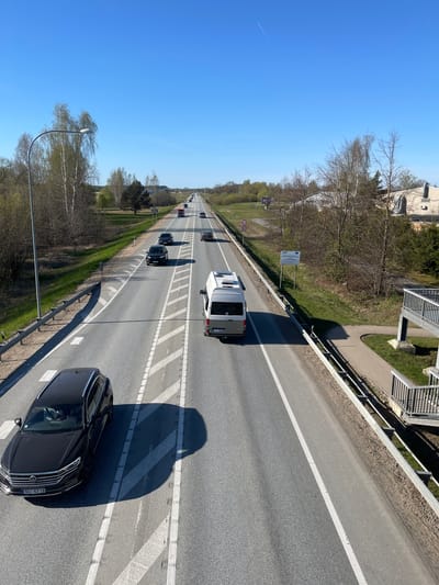 The image shows a highway with multiple lanes in Ādaži, Latvia. Several vehicles are traveling on the road, including a black Volkswagen car in the foreground, a white van, and other cars further down the highway. The road has white lines marking the lanes and a guardrail on the side. Trees line the side of the highway. The sky is clear and blue. A pedestrian overpass is visible on the right side of the image. A street lamp is visible on the left side of the image.