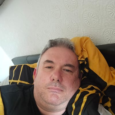A middle-aged man with grey hair and light stubble is depicted lying down indoors in Holywell, United Kingdom. He is looking directly towards the camera with a neutral or slightly contemplative expression. He is partially covered by or wearing a black garment featuring prominent yellow stripes and patterns, suggesting it could be a jacket, blanket, or similar attire. Behind his head, a dark, possibly leather, surface is visible, likely a headrest or cushion. The dominant background is a white, textured surface, possibly wallpaper or a bedspread, adorned with a repeating embossed pattern of stylized flowers or stars enclosed within circles. The lighting is bright and diffused, indicating an indoor setting with ample illumination. No text is visible in the image.