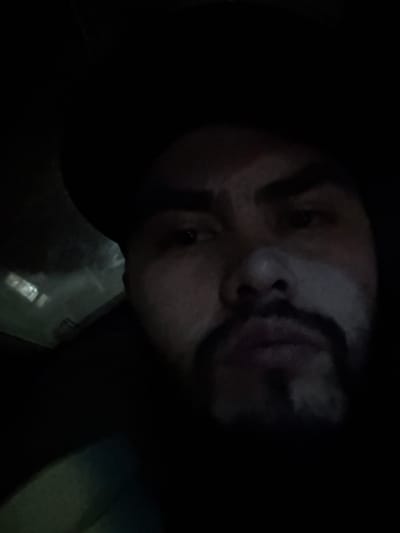 A person's face is tilted and shadowed. The person has a dark beard and their expression is difficult to discern due to the low lighting. Hints of what may be a glass or plastic container are visible near the bottom edge of the image, adding context without clarifying the overall scene. The focus is primarily on the upper half of the face, with the lower portion obscured.