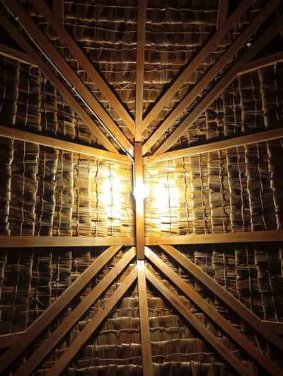 A low-angle shot depicts the interior of a structure with a thatched roof, likely in Namplohbaro, Indonesia. The roof is constructed from straw-like material laid in horizontal rows, secured by darker twine. Wooden beams form a radial pattern, converging at a central point where a light fixture is positioned. The light casts a bright glow, illuminating the surrounding area and creating stark shadows. The overall impression is one of rustic construction, with a focus on the interplay of light and shadow within the architectural framework.