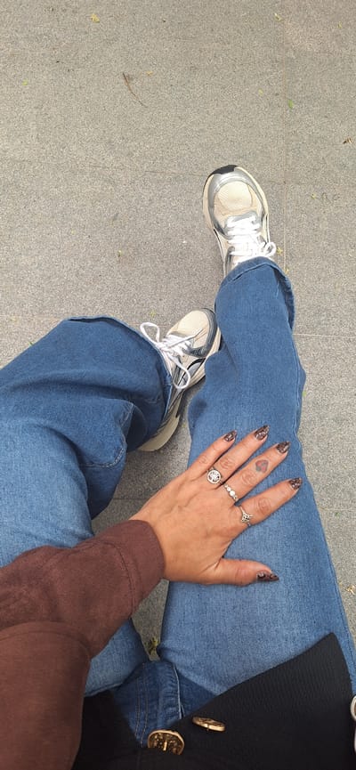 The image is a high-angle, close-up shot from a first-person perspective, featuring a person's legs, right hand, and elements of their clothing. The person is wearing loose-fitting blue denim jeans and white and silver sneakers with white laces, resting on a gray, speckled paved surface. Small pieces of dried leaves and debris are scattered across the textured, tiled ground.
The person's right hand, with an animal print (likely leopard print) nail design, rests casually on their left thigh. Several rings adorn the fingers: a band with three small stones on the middle finger, a larger circular ring with a patterned design on the index finger, and a ring with a quatrefoil-like shape on the pinky finger. A small, dark red heart tattoo is visible on the ring finger.
Part of a brown, long-sleeved garment, likely a top or sweater, is visible on the right arm. Further to the upper right, a section of a black textured garment, possibly a coat or jacket, is shown, featuring two prominent gold-colored decorative buttons.
The scene takes place outdoors during bright daylight. The ground appears to be a public walkway or square, consistent with an urban or suburban setting. The image primarily captures a casual, relaxed pose, highlighting details of personal style. No visible text or specific location cues are present within the image itself, though the provided context suggests Santa Cruz de Tenerife, Spain.