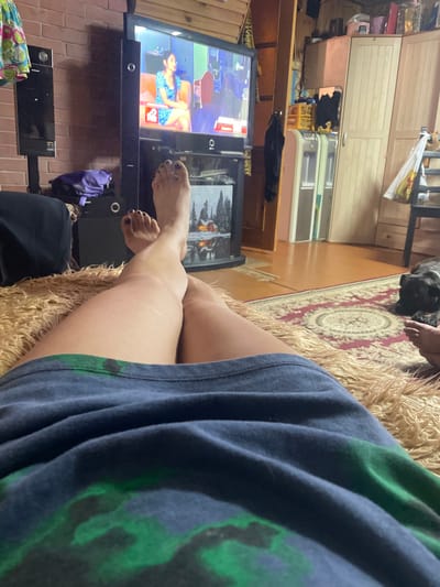 The image depicts a person's legs and torso, draped in a blue and green patterned garment, resting on a textured, tan couch. The person's bare feet, with dark nail polish, are visible. To the left, a television screen displays a woman in a blue dress sitting on an orange couch. The background reveals a room with wooden paneling, a rug with a circular pattern, and a dog lying down in the distance. Storage units with drawers are also visible. The scene appears to be indoors, likely in a home environment. The overall mood is relaxed and casual, suggesting a person watching television. There is no information about the time of day or weather. No visible text is clearly legible.