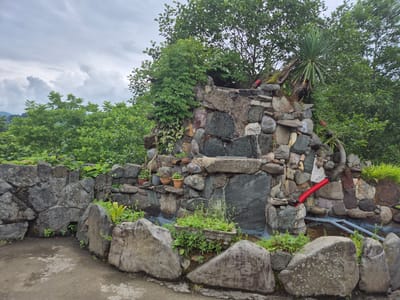 This is an eye-level, medium shot of a man-made rock waterfall in Charnali, Georgia. The waterfall is constructed from various sizes and colors of stones, with several plants and small potted plants growing on and around it. A red pipe is visible emerging from the top right of the waterfall, and grey pipes are visible at the bottom right. The background consists of lush green trees and foliage under a cloudy sky. In the foreground, a concrete path leads towards the waterfall. The overall scene depicts a garden or park area with a decorative water feature.
