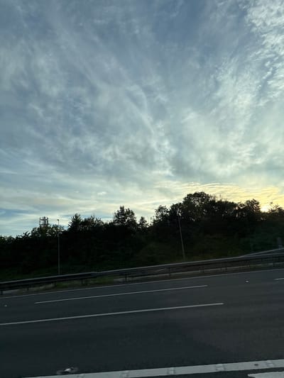 The image shows a rural landscape, likely in South Staffordshire, United Kingdom. A highway runs through the foreground, with lanes marked by white lines. Beyond the highway is a dense line of dark green trees, with a single street light visible amidst them. In the distance, to the left, a tall, metal telecommunications tower stands above the treeline. The sky above is filled with wispy, white clouds against a pale blue background. The lighting suggests it is either early morning or late afternoon, with a hint of golden light on the right side of the horizon. There are no people or vehicles visible in the image. The overall impression is one of natural beauty combined with infrastructure.